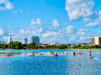 Paddleboarders and kayakers enjoying a sunny urban lake with downtown skyline, green shoreline, and blue sky dotted with puffy clouds