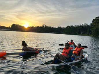 Four people paddling clear-hull kayaks on a calm lake at sunset, wearing bright orange life jackets with a tree-lined shore in the background