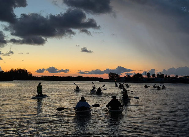 Group of silhouetted kayakers and a paddleboarder crossing a calm coastal inlet at sunset, orange-blue sky with dramatic clouds and a tree-lined shoreline.