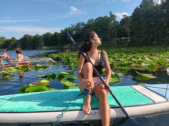 Woman paddleboarding on a turquoise board through green water lilies on a sunny lake with a tree-lined shore and blue sky.