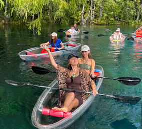 Friends in a transparent kayak posing and paddling on crystal-clear turquoise spring water beneath hanging green trees, with other kayakers nearby.
