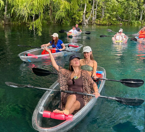 Friends in a transparent kayak posing and paddling on crystal-clear turquoise spring water beneath hanging green trees, with other kayakers nearby.