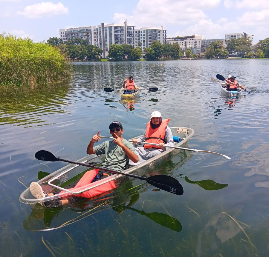 Two people in a clear see-through kayak wearing orange life jackets paddle on an urban lake near reeds, with two other kayakers and waterfront apartment buildings in the background.