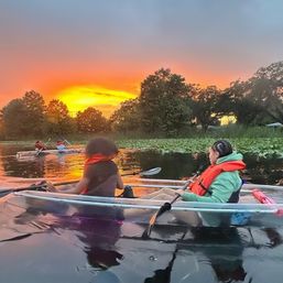 Two paddlers in life jackets glide in a clear kayak across a calm lake at sunset, orange sky and tree-lined shore reflected on the water near lily pads — scenic sunset kayaking.