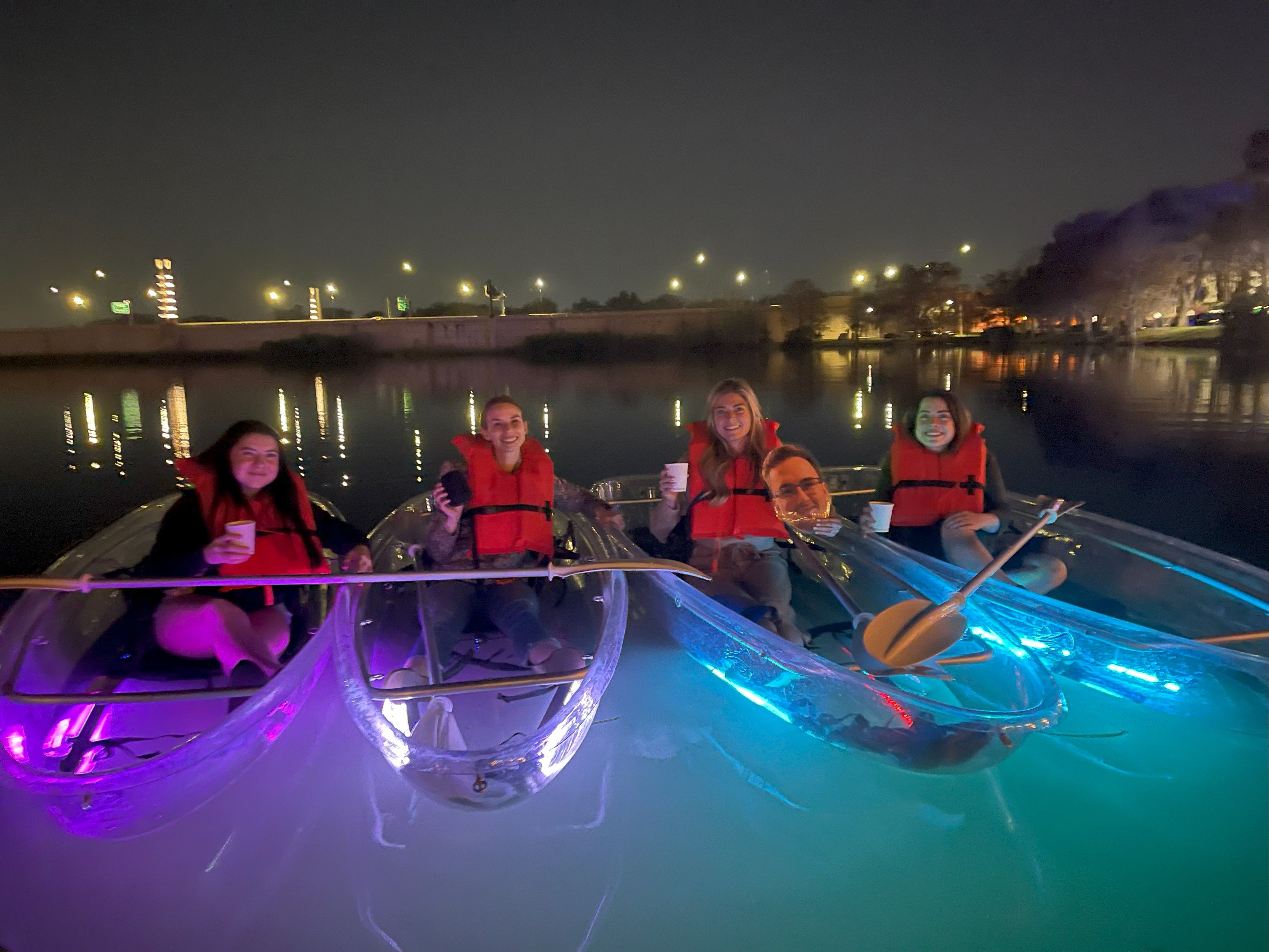 Five people in clear LED-lit kayaks at night on a calm urban river, wearing red life jackets and holding cups with city lights reflecting on the water.