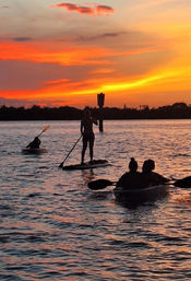 Silhouetted paddlers — one on a stand-up paddleboard and two in kayaks — gliding across a calm bay at a vibrant orange-pink sunset with shimmering water reflections.