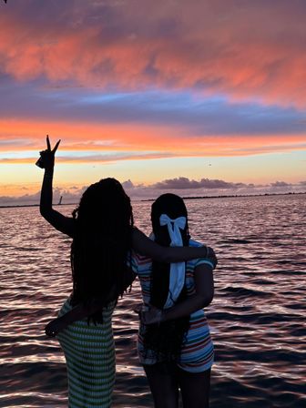 Silhouettes of two friends embracing by the ocean at sunset, vibrant pink-orange clouds and rippling water reflecting the colorful sky, one friend flashing a peace sign.