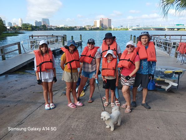 Group of eight people in orange life jackets and a small white dog on a lakeside dock with kayaks and an urban skyline across the water.