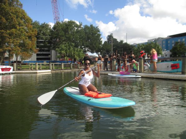 Smiling woman in a white 'BRIDE' swimsuit kneeling on a turquoise stand-up paddleboard near a wooden dock, holding a paddle with a group of people and extra boards on the urban waterfront under a sunny sky.