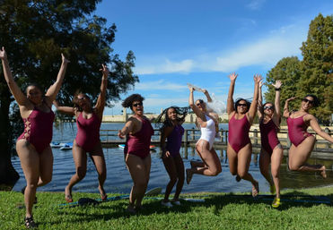 Lakeside summer group photo of eight women in maroon and white swimsuits jumping and cheering on a sunny waterfront lawn with a wooden dock and blue sky.