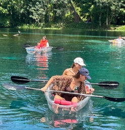 Two people paddling a clear-bottom kayak on a crystal-clear spring-fed river with lush green shoreline and a family in life vests in the background