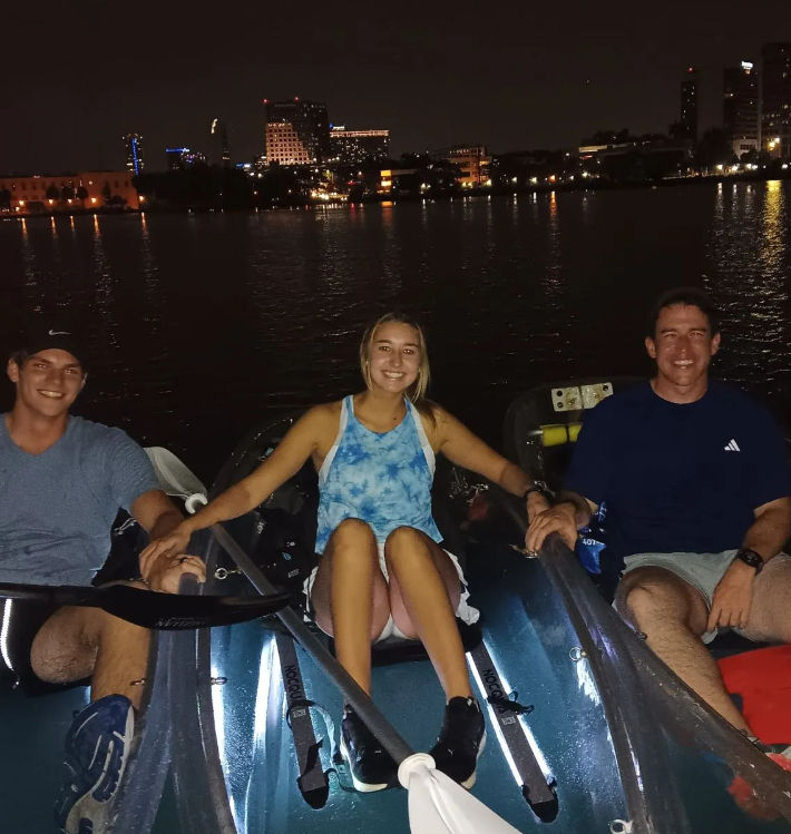 Night kayaking: three smiling people seated in kayaks on a calm river, holding paddles and hands, with a lit urban skyline reflected across the water.