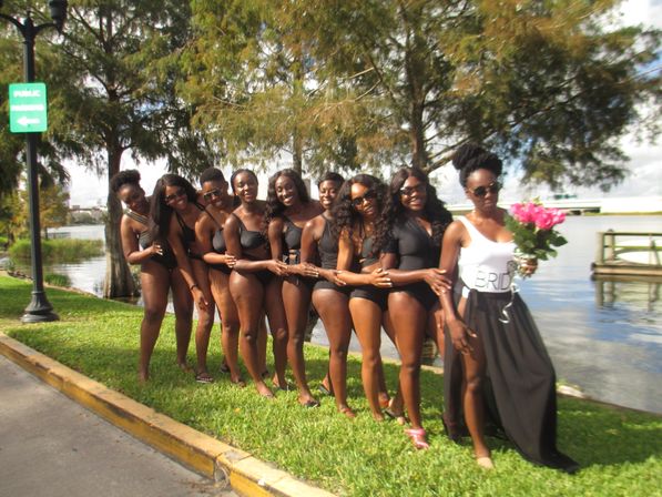 Lakeside bridal party: nine women in black swimsuits lined up arm-in-arm, bride in white 'BRIDE' top and black skirt holding pink bouquet by trees and a small dock