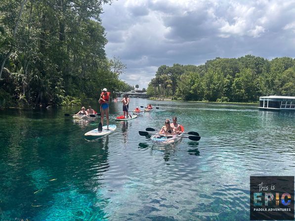 Paddleboarders and kayakers enjoying a crystal-clear turquoise Florida spring river with lush tree-lined banks and a cloudy sky — outdoor paddling adventure.