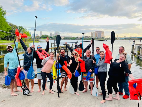 Energetic group of adults on a dock holding paddles and bright red life jackets, ready for paddleboarding at an urban waterfront with a downtown skyline in the background.