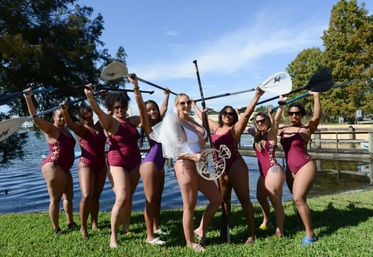 Cheerful bachelorette party by a lake — bride-to-be in white swimsuit and veil with bridesmaids in matching burgundy swimsuits holding stand-up paddleboard paddles overhead on a sunny waterfront lawn