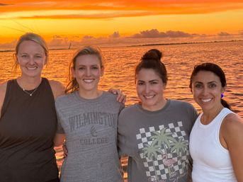 Four friends smiling arm-in-arm at a waterfront sunset, vivid orange sky reflecting on calm water with a distant bridge on the horizon.