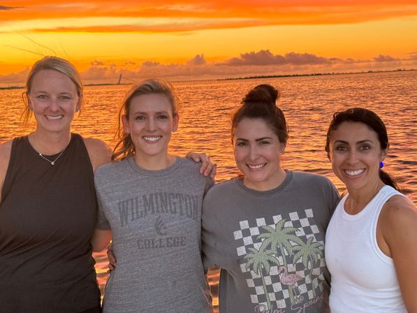 Four friends smiling arm-in-arm at a waterfront sunset, vivid orange sky reflecting on calm water with a distant bridge on the horizon.