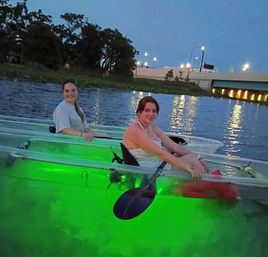 Two kayakers in clear kayaks glowing green from underwater lights, paddling on a river at dusk with a lit bridge and tree-lined shore reflected in the water.