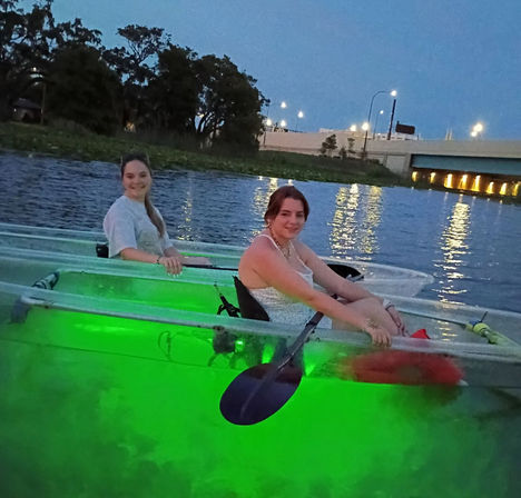 Two kayakers in clear kayaks glowing green from underwater lights, paddling on a river at dusk with a lit bridge and tree-lined shore reflected in the water.