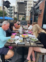Adults at a long outdoor patio table on an urban restaurant terrace taking a calligraphy workshop, writing on worksheets with pens while eating salads and sipping drinks under string lights and city apartments in the background.