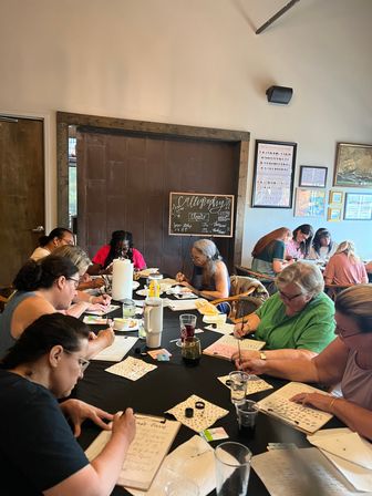 Indoor modern calligraphy workshop: adults seated around a long table in a bright studio practicing lettering with nib pens, ink pots, practice sheets and drinks.