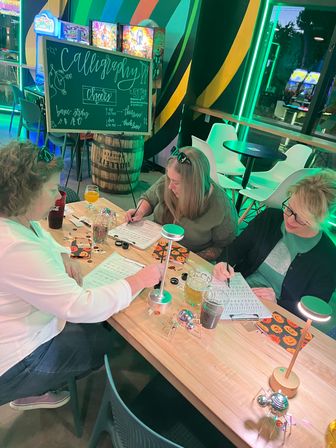Three people practicing modern calligraphy at a wooden table in a neon-lit urban cafe workshop, with a chalkboard reading “Calligraphy,” practice sheets, pens, drinks, small lamps and Halloween-themed napkins.