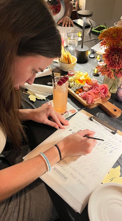 Person practicing modern calligraphy at a cozy indoor workshop table, tracing strokes on a worksheet beside a cocktail with raspberries, a wooden charcuterie board, autumn flowers and candlelight.