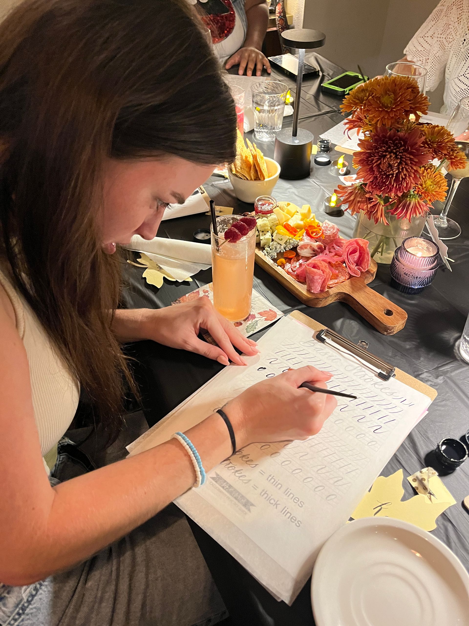 Person practicing modern calligraphy at a cozy indoor workshop table, tracing strokes on a worksheet beside a cocktail with raspberries, a wooden charcuterie board, autumn flowers and candlelight.