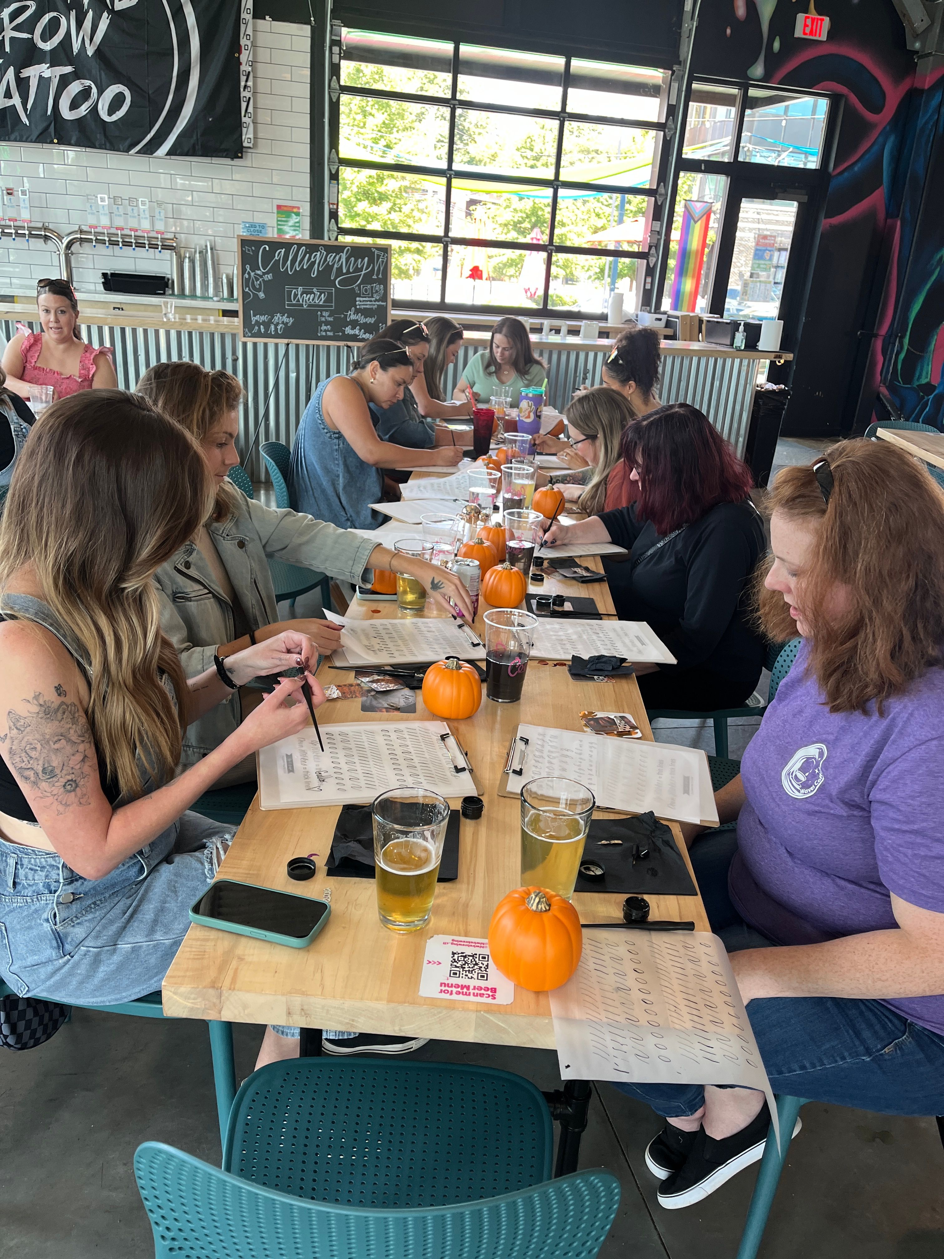 Group of adults at a long wooden table in an industrial taproom doing a calligraphy workshop, worksheets, small pumpkins and pints of beer with large garage windows and mural in the background