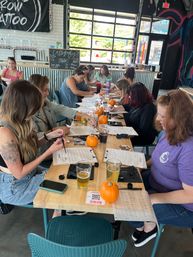 Group of adults at a long wooden table in an industrial taproom doing a calligraphy workshop, worksheets, small pumpkins and pints of beer with large garage windows and mural in the background