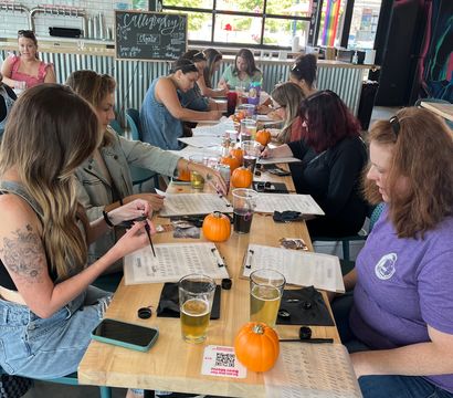 Group of adults at a long wooden table in an industrial taproom doing a calligraphy workshop, worksheets, small pumpkins and pints of beer with large garage windows and mural in the background