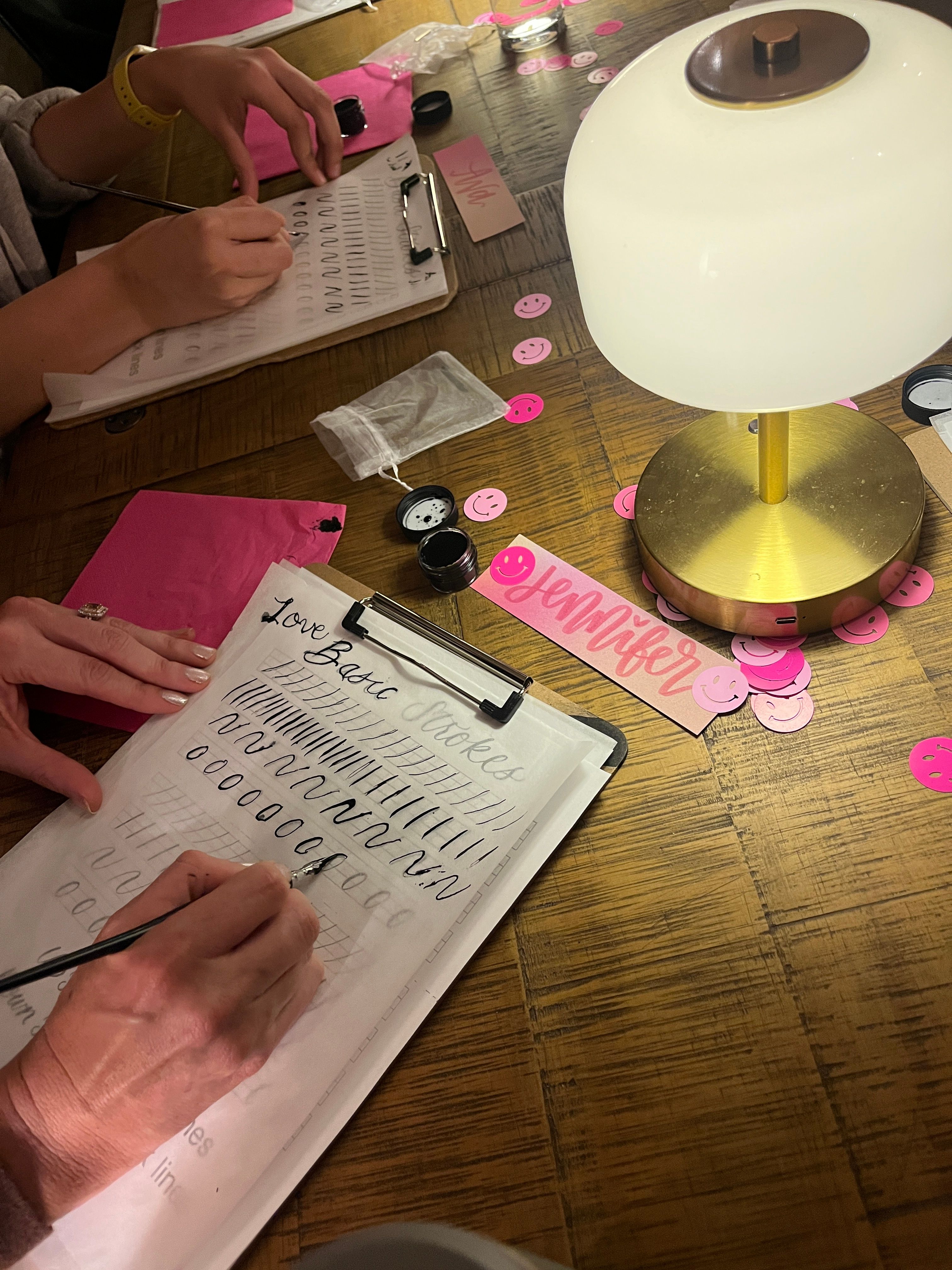 Close-up of hands practicing brush lettering at a wooden table during a cozy calligraphy workshop, with ink pots, practice sheets on clipboards, pink smiley confetti, a brass desk lamp and a pink name tag