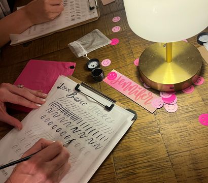 Close-up of hands practicing brush lettering at a wooden table during a cozy calligraphy workshop, with ink pots, practice sheets on clipboards, pink smiley confetti, a brass desk lamp and a pink name tag