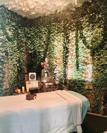 Cozy spa treatment room with a massage table draped in white linens and a green throw, surrounded by a faux stone wall covered in hanging ivy, candle accents, and soft cloud-like ceiling decor for a relaxing indoor garden vibe.