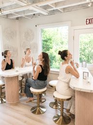 Bright, light-filled beauty bar interior with four smiling women seated on gold-accented stools at a white marble counter, testing perfume and makeup near a large window with lush green view.