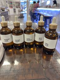 Five amber glass dropper bottles of fragrance oils labeled 'floriental wood', 'aromatic aquatic', 'earthy patchouli', 'green fruity', and 'clean musk' lined up on a wooden table with a bright, blurred boutique interior and counter in the background.