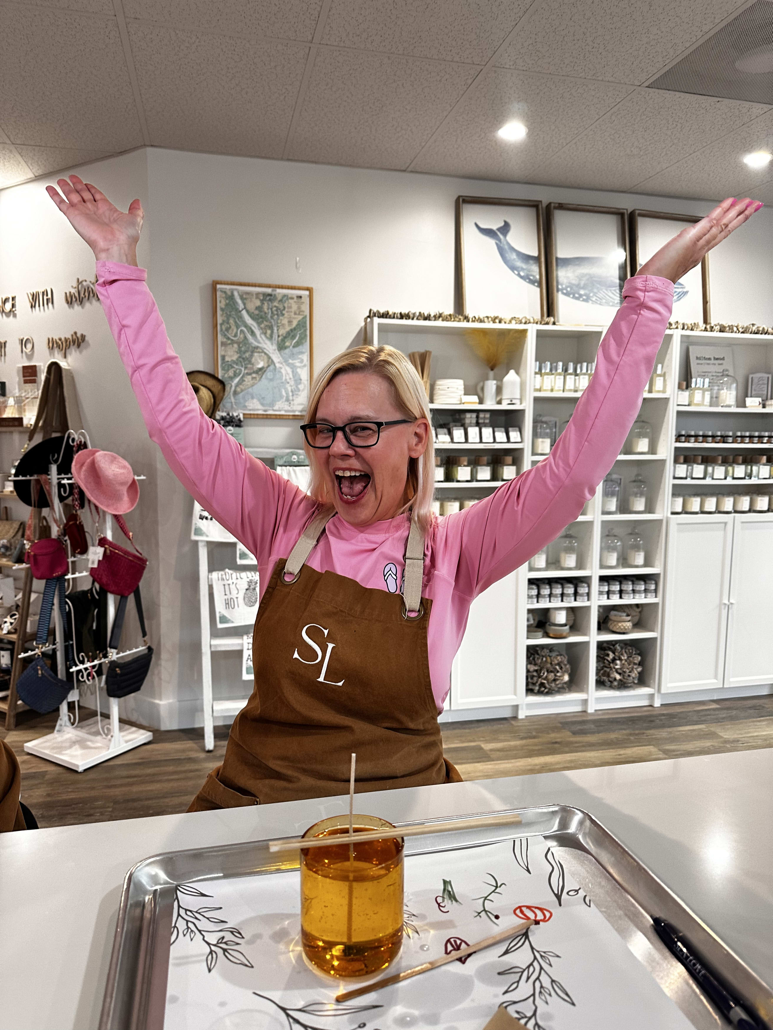 Cheerful shop attendant in a pink shirt and brown apron raises both arms in a boutique candle-making studio, shelves of jars and home goods behind and a glass candle jar with a wick on a tray in the foreground.