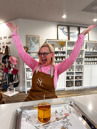 Cheerful shop attendant in a pink shirt and brown apron raises both arms in a boutique candle-making studio, shelves of jars and home goods behind and a glass candle jar with a wick on a tray in the foreground.