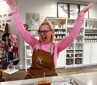 Cheerful person in a brown apron raising both arms in an airy artisan candle workshop, smiling over a jar of amber poured wax on a tray with shelves of candle supplies and decor in the background.