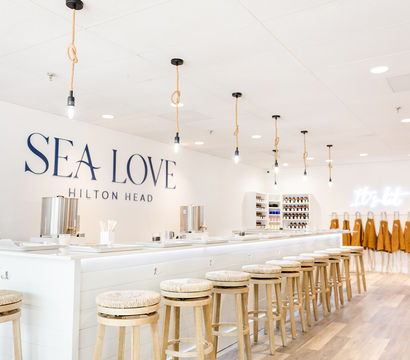 Bright coastal-themed studio interior in Hilton Head with a long white counter, row of woven-seat wooden barstools, rope-hung pendant lights, and aprons and supplies along the back wall.