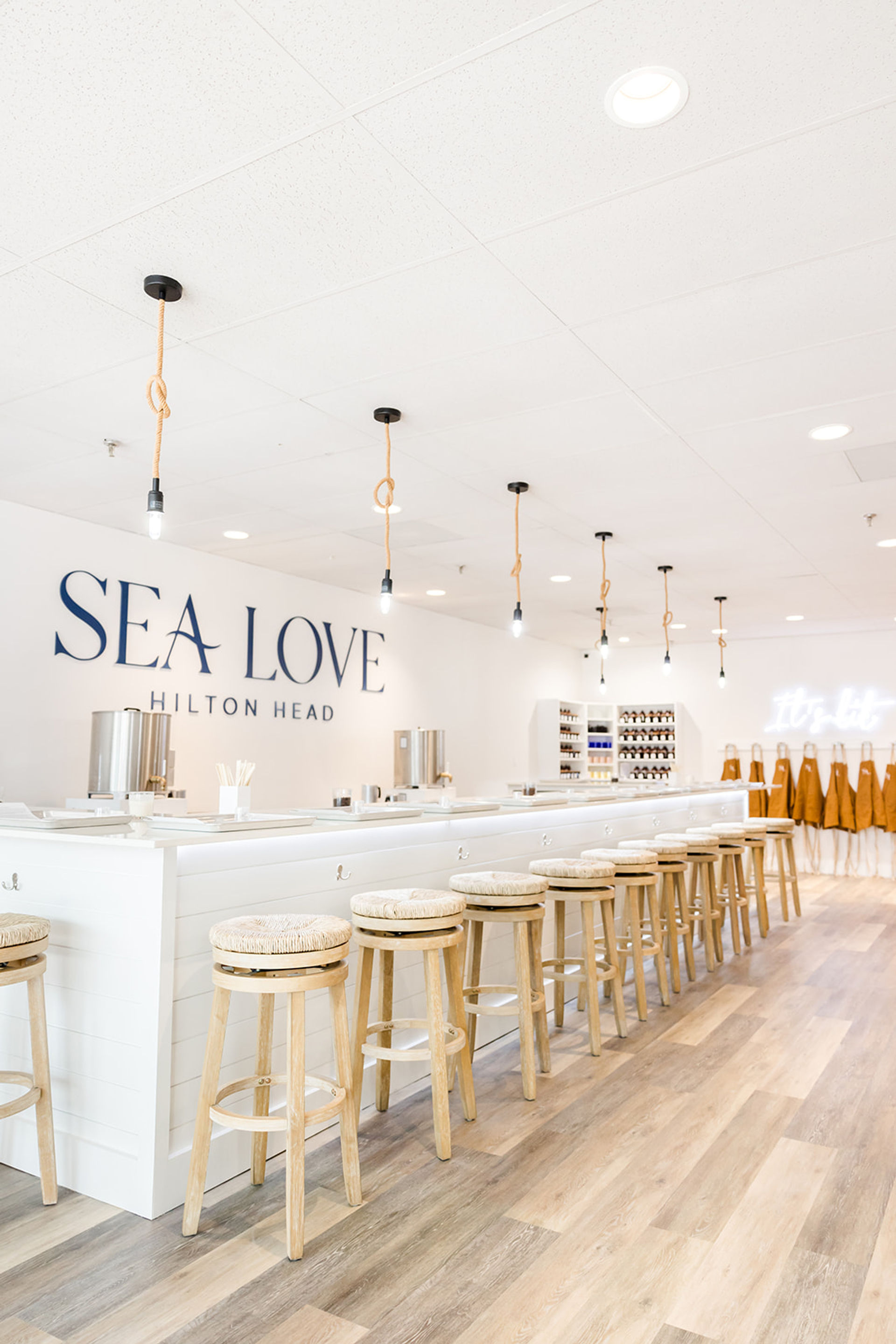 Bright coastal-themed studio interior in Hilton Head with a long white counter, row of woven-seat wooden barstools, rope-hung pendant lights, and aprons and supplies along the back wall.