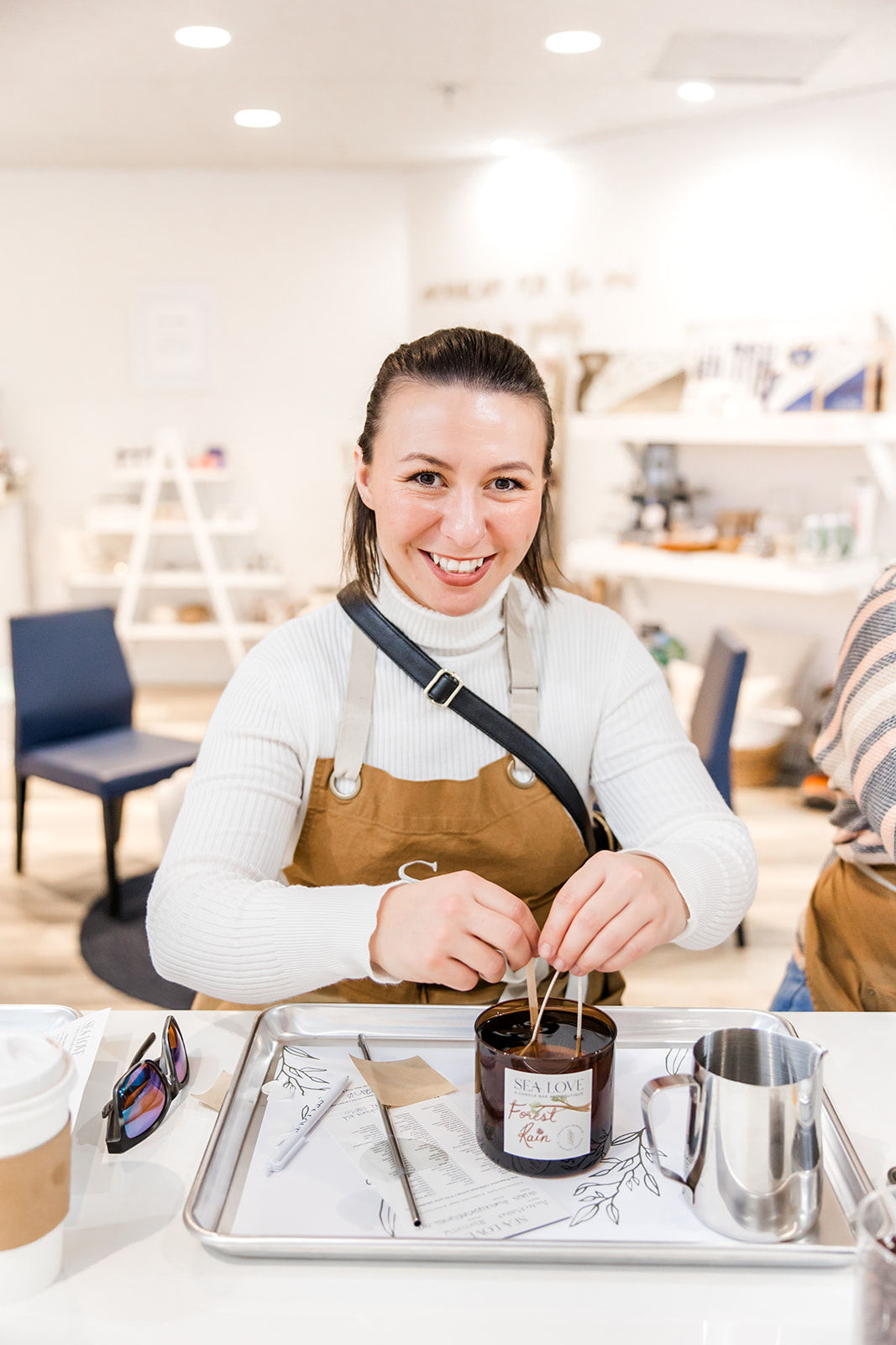 Smiling woman at a DIY candle-making workshop in a bright craft studio, inserting wicks into a glass candle jar on a tray with tools, coffee cup, and metal pitcher.