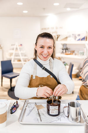 Smiling woman at a DIY candle-making workshop in a bright craft studio, inserting wicks into a glass candle jar on a tray with tools, coffee cup, and metal pitcher.