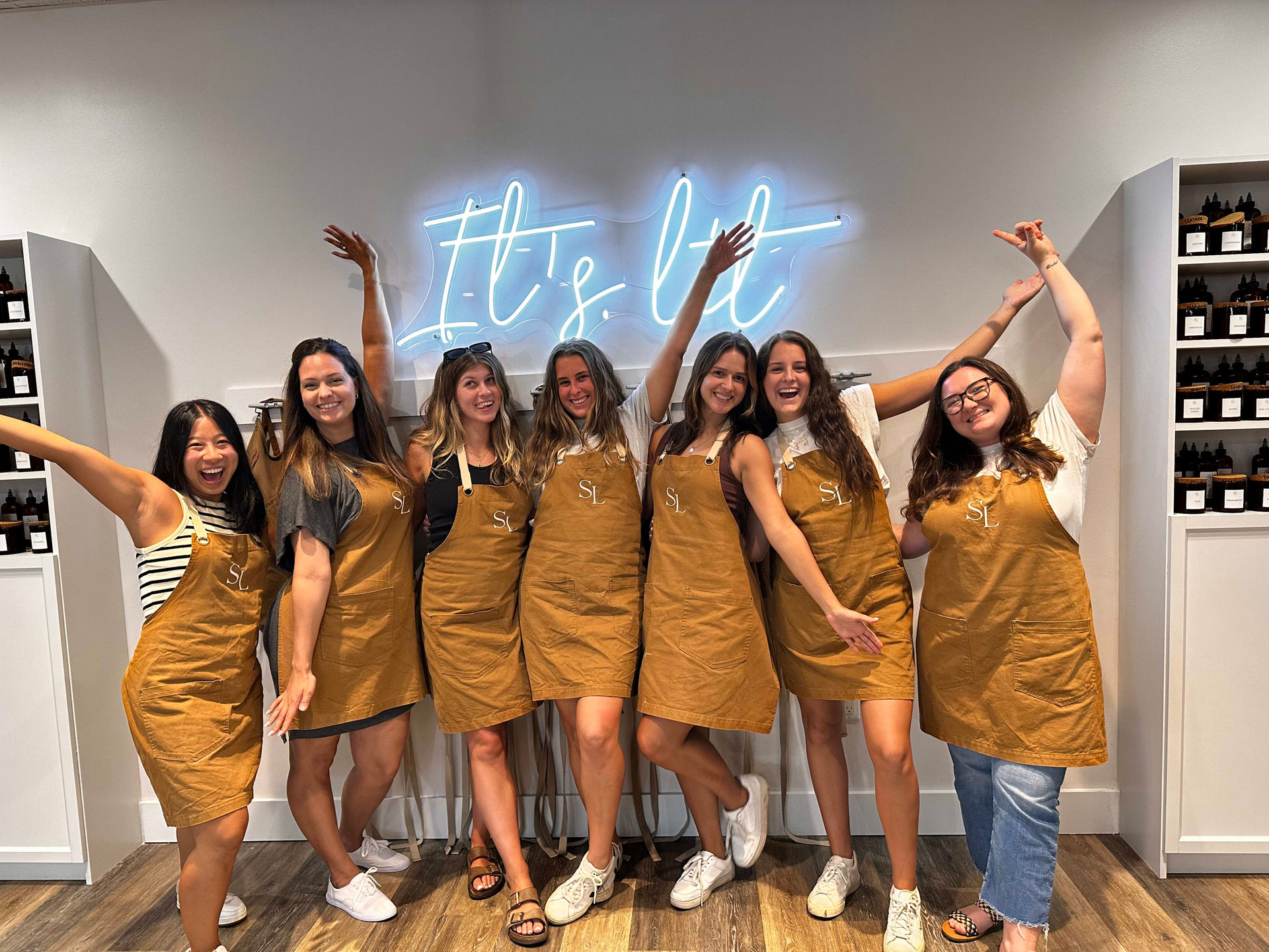 Seven smiling people in matching mustard-brown aprons pose energetically with arms raised under a blue neon 'It's lit' sign in a bright boutique studio with wood floors and shelves of amber bottles.