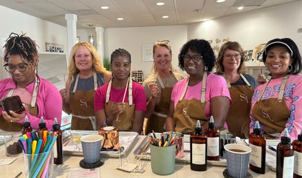Seven women in aprons at a bright craft studio candle-making workshop, smiling behind a counter filled with copper candle tins, fragrance bottles labeled Cucumber/Lavender/White Tea, colored pencils and coffee cups.