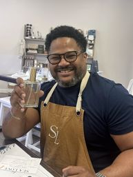 Smiling man wearing glasses and a brown apron holds a glass perfume spray bottle in an artisanal scent workshop with shelves of candles and supplies in the background.