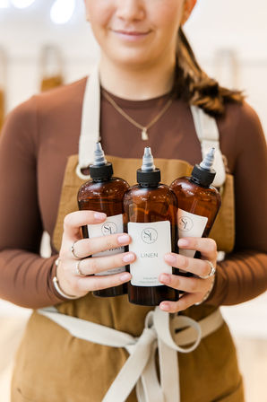 Close-up of a person in an apron holding three amber linen fragrance refill bottles with nozzle tops in a cozy boutique