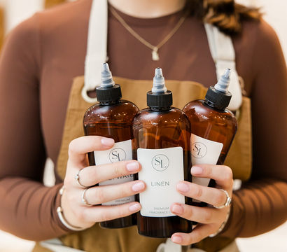 Close-up of a person in an apron holding three amber linen fragrance refill bottles with nozzle tops in a cozy boutique