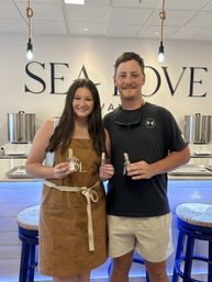 Smiling pair holding small perfume bottles in a bright seaside-inspired shop with rope pendant lights, blue stools and stainless mixing kettles in the background.